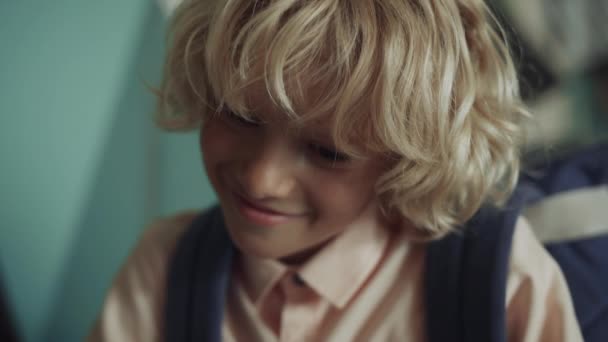 Boldog szőke fiú szórakozik az iskolai szünetben ülő folyosón közelről. Vidám mosolygós fickó, aki lenéz játszani a barátaival. Portré vidám iskolás fiú hátizsák ellenőrző osztálytársak. - Felvétel, videó