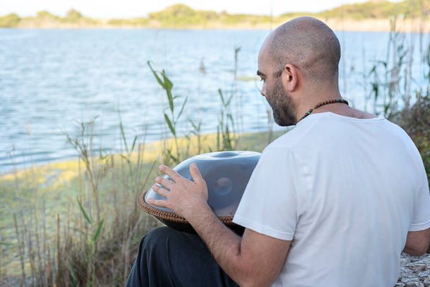 Rückansicht eines Yogi-Mannes, der eine traditionelle Stahlhandpfanne spielt, am Ufer des Sees sitzend, beruhigende Musik - Foto, Bild