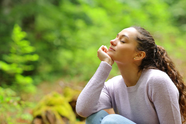 Relaxed woman sitting with closed eyes breathing fresh air in a beautiful green forest - Фото, зображення
