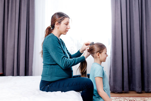 Charming little girl is smiling while her beautiful young pregnant mother is combing daughters hair, side view. photo of young woman using brush combing little girl hair. - 写真・画像