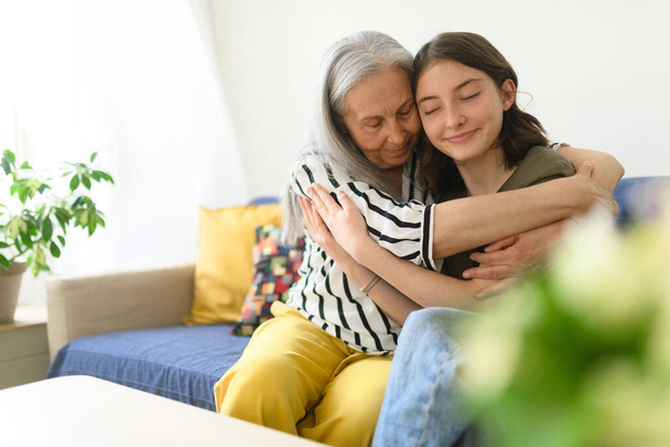A senior grandmother with teenage granddaguhter having good time together at home, hugging. - Фото, изображение