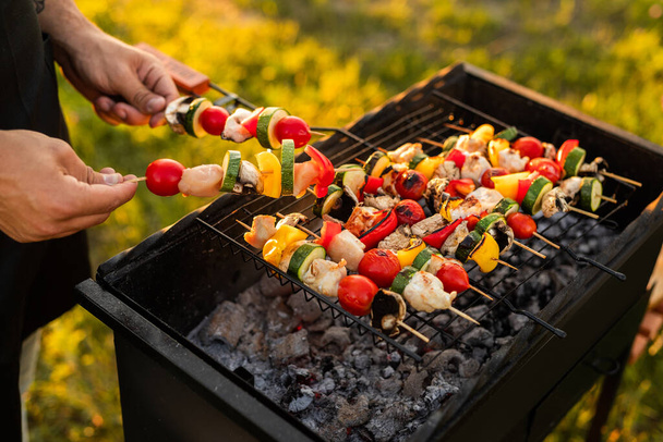From above anonymous man holding raw chicken and vegetable skewers over grill with charcoal during barbecue on sunny weekend day - Фото, изображение
