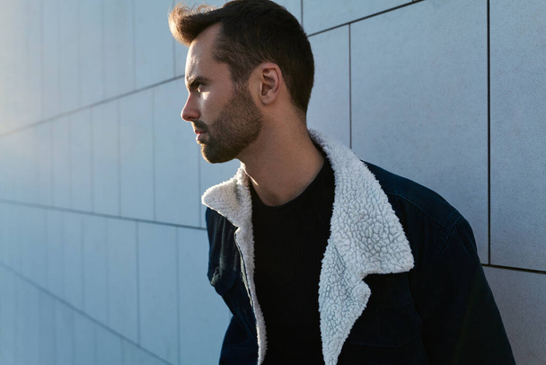 Concentrated young bearded ethnic male with dark hair in stylish casual clothes standing near tiled wall and looking away on sunny day in city - Photo, Image