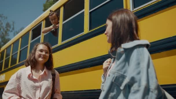 Deux jolies filles qui parlent dans le bus scolaire jaune. Mignons camarades de classe souriants bavardant debout près d'un autobus scolaire au soleil. Curieux écolier regardant par la fenêtre observant des amis adolescents. Concept d'étude. - Séquence, vidéo