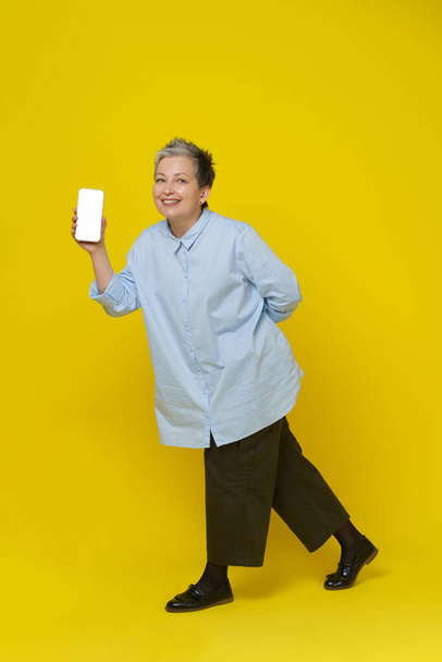 Showing phone in hand mature grey haired woman smiling on camera. Pretty woman in blue shirt and brown trousers isolated on yellow background. Mobile app advertising. Mockup product placement. - 写真・画像