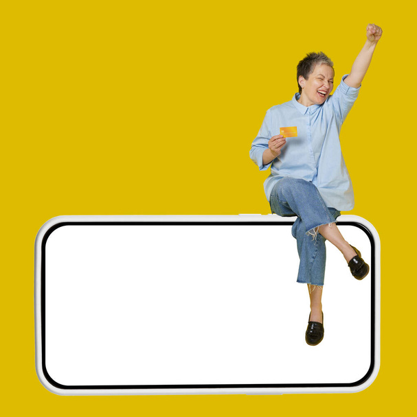 Excited, happy mature grey haired woman sitting on a giant, huge smartphone, in casual blue shirt and jeans with hand raised up in a win, isolated on yellow background.  - Фото, изображение