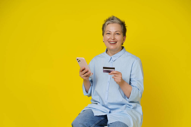 Grey haired woman hold phone and debit or credit card in hand making online payment or shopping. Pretty mature woman in blue shirt and jeans isolated on yellow background. - Photo, image