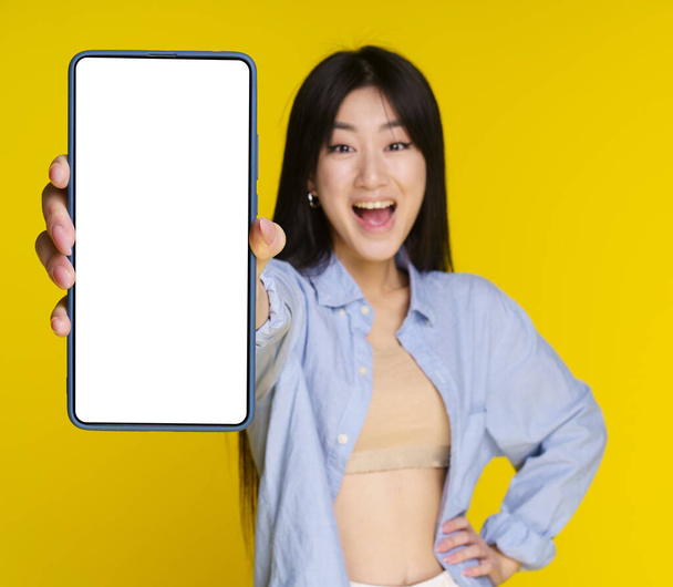Young asian girl holding smartphone in hand showing white screen for your mock up mobile app advertisement and excited smile on camera isolated on yellow background. Selective focus on phone. - Photo, Image