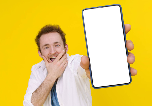 Cant believe, shocked young man with smartphone showing a white empty screen isolated over yellow game, bet, lottery win. Product placement. Mobile App Advertisement.  - Фото, изображение