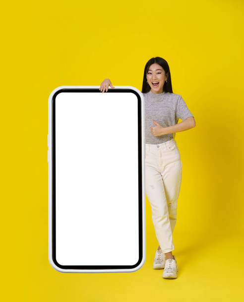 Bájos ázsiai lány gesztikuláló hüvelykujj fel állni közelében hatalmas, hatalmas okostelefon fehér képernyőn boldog öltözött alkalmi elszigetelt sárga háttér. A szabad tér gúnyolódik. Termék elhelyezése mobil alkalmazás hozzáadás. - Fotó, kép