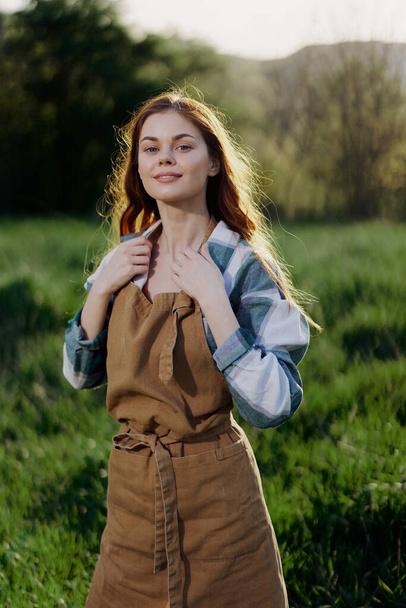A woman gardener in an apron stands in a field of green grass outdoors, smiling on a summer day into a sunset. High quality photo - Photo, Image