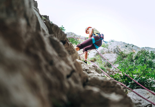 Active climber woman in protective helmet abseiling from cliff rock wall using rope with belay device and climbing harness. Active extreme sports time spending concept. - Фото, зображення