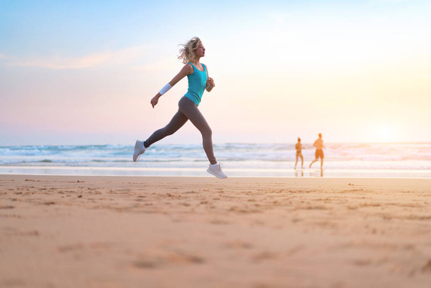 Active sporty woman run along ocean surf by water pool to keep fit and health. Sunset sand beach background with sun. Woman fitness, jogging workout and sport activity on summer family holiday. - Photo, image