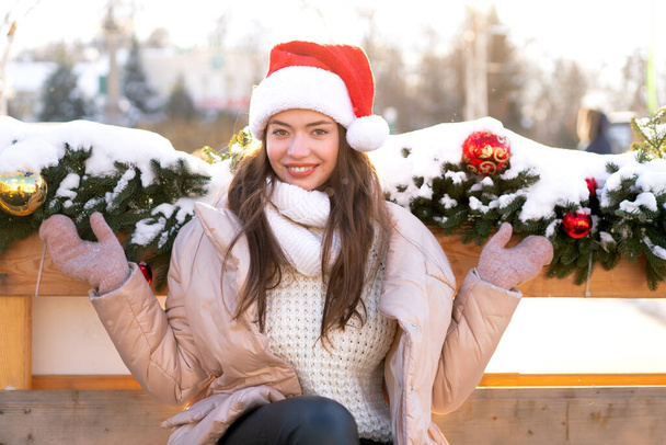 Winter portrait young adult beautiful woman in Santa hat. Christmas mood. Snowing. Winter beauty fashion concept. Pretty young woman sitting bench outdoor near Christmas garland - Photo, Image