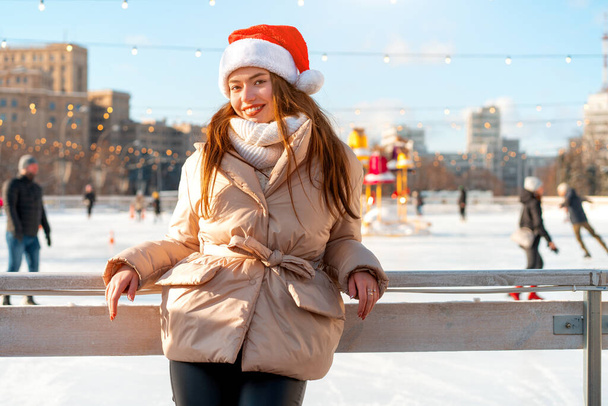 Outdoor portrait young beautiful fashionable woman wearing Santa hat, posing street of European city. Winter Christmas holidays concept. ice skate rink background. Lean on fence near ice skate rink - Fotó, kép