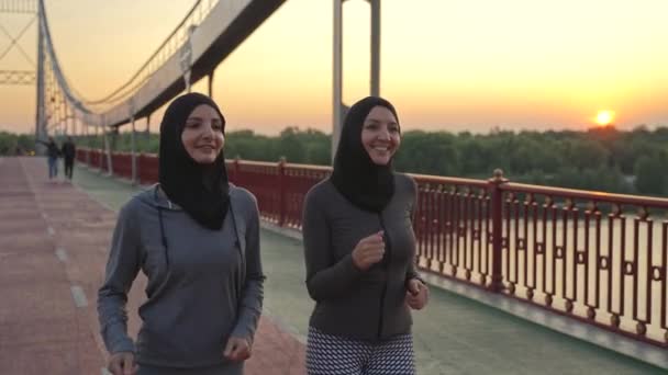 Hembras sonrientes en hiyabs y ropa deportiva corriendo en la pasarela cruzando el río de la ciudad al amanecer. Deportivos corredores árabes disfrutando de ejercicio conjunto por la mañana, activo saludable estilo de vida deportivo urbano concepto - Imágenes, Vídeo