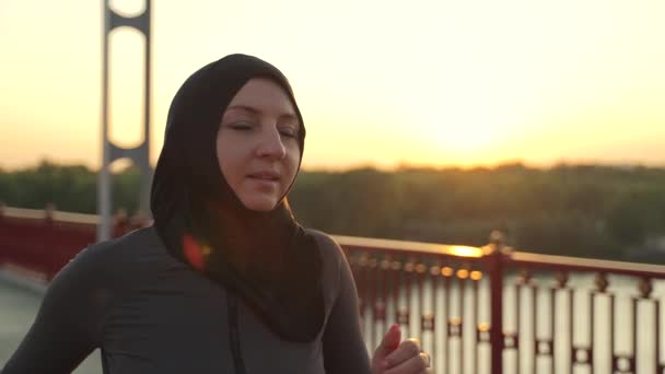 Κοντινό πλάνο μιας γυναίκας Άραβας με χιτζάμπ να τρέχει στην γέφυρα της πόλης νωρίς το πρωί. Ενεργός υγιής δρομέας απολαμβάνοντας καρδιο προπόνηση στη φύση, σπορ τρόπο ζωής έννοια - Πλάνα, βίντεο