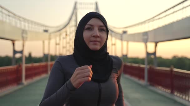 Κοντινό πλάνο του ενεργού αθλητικού muslim θηλυκό φορώντας hijab τζόκινγκ το πρωί. Ενηλίκων ταιριάζει γυναίκα κάνει σπορ σε εξωτερικούς χώρους, τρέχει σε όλη την πόλη footbridge, υγιές χόμπι, ενεργός τρόπος ζωής έννοια - Πλάνα, βίντεο