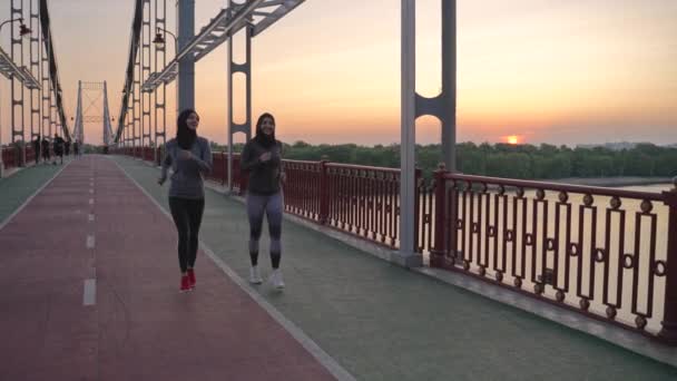 Две здоровые арабские женщины в хиджабах и спортивной одежде наслаждаются вечерней пробежкой по городскому пешеходному мосту. Активные мусульманские бегуны проводят свободное время, занимаясь кардио-тренировками на открытом воздухе - Кадры, видео