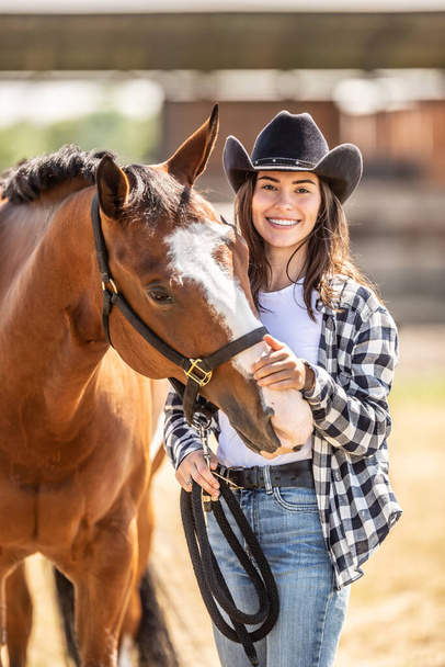 Przystojna młoda kobieta w kowbojskim kapeluszu głaszcze konia na kotlety uśmiechając się do kamery. - Zdjęcie, obraz
