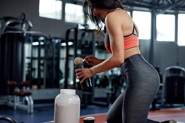 Una deportista en forma está mezclando proteína en polvo y haciendo una bebida proteica en un gimnasio. - Foto, Imagen