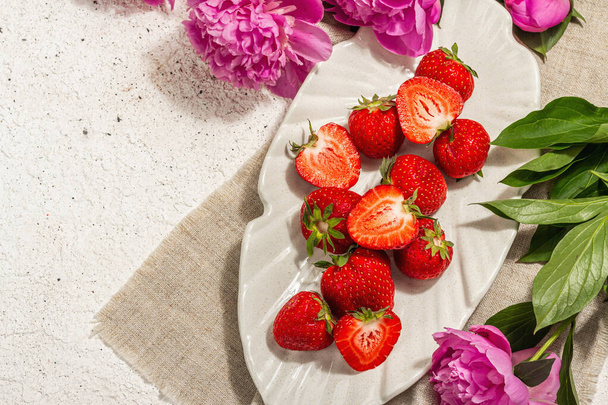 Sweet ripe strawberries and fragrant peonies bouquet. Summer plaster background, seasonal design, hard light, dark shadow, top view - Foto, Bild