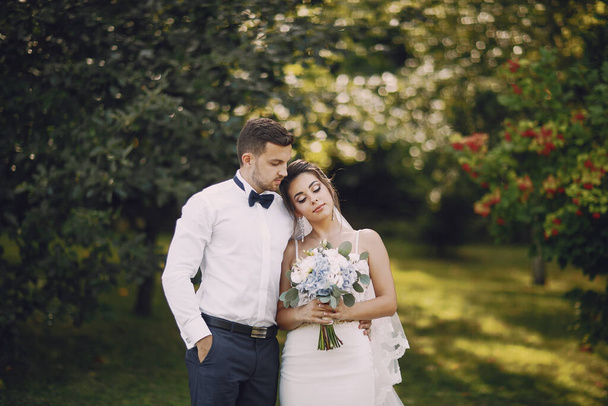 A young and beautiful bride and her husband is standing in a summer park with bouquet of flowers - Φωτογραφία, εικόνα