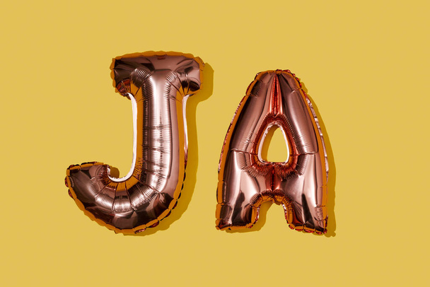 ドイツ語、オランダ語、デンマーク語などのドイツ語で黄色の背景に「 ja 」という言葉を形成する金属製のピンク色の手紙型の風船があります - 写真・画像