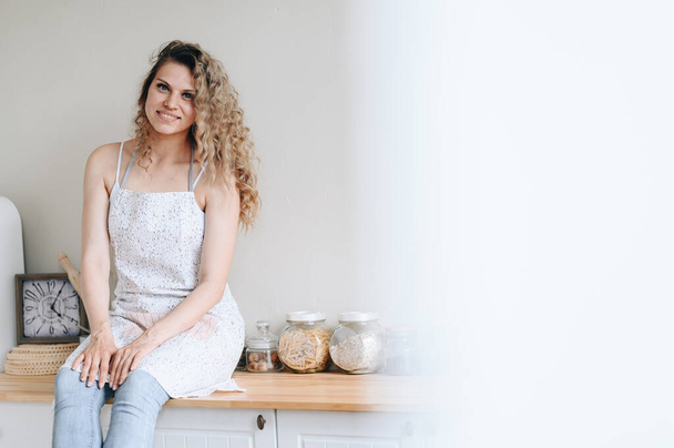 Young beautiful cute curly woman is sitting on table in kitchen in an apron. concept of young housewife is preparing to eat. on banner. copy space - Φωτογραφία, εικόνα