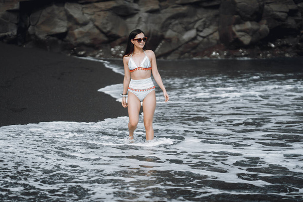 Brunette girl walking on the black sand beach wearing a cute bathingsuit in Bali Indonesia - Foto, imagen