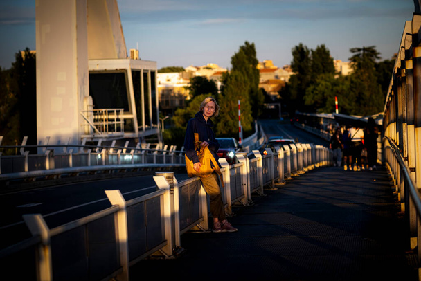 Una turista donna con uno zaino giallo si erge su un ponte durante il tramonto. - Foto, immagini