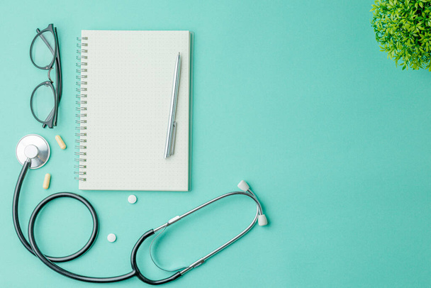 Open notebook with a pen and glasses on a green hospital background flat lay. top view. Nurse workplace concept. A stethoscope and pills on the doctor work desk. - Photo, Image