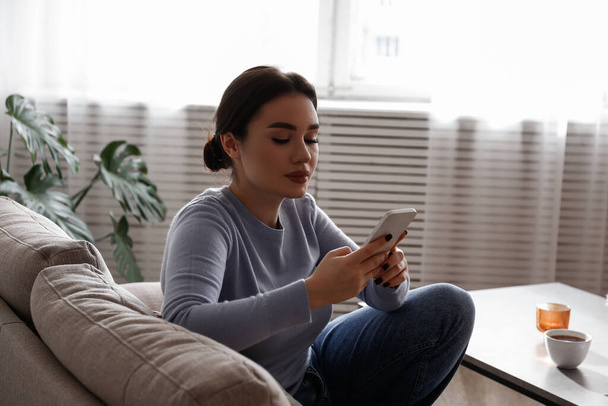 Portrait of young brunette woman with concentrated facial expression sitting on the couch typing on the phone. Joyful female having a video call. Background, copy space, close up. - Fotoğraf, Görsel