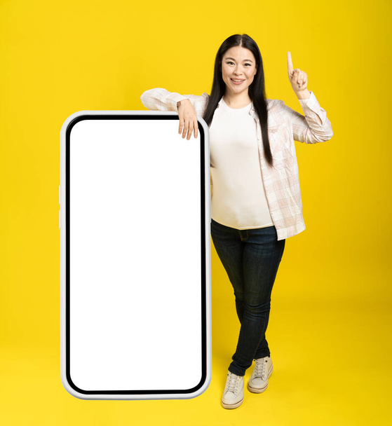 Asian woman pointing finger one as attention leaned back on giant, huge smartphone with blank white screen, wearing casual isolated on yellow background. Free space mock up. - Valokuva, kuva