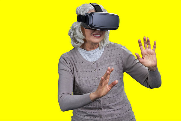Old happy caucasian woman wearing virtual reality goggles. Senior lady playing on color background. - 写真・画像