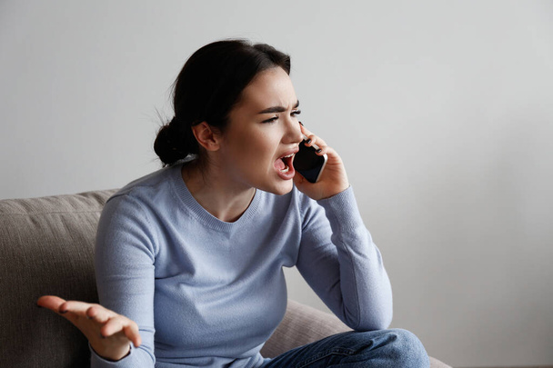 Portrait of irritated young woman arguing on phone. Outraged female talking angrily, shouting at cellphone. Customer support frustration concept. Copy space for text, white wall background, close up. - Photo, image