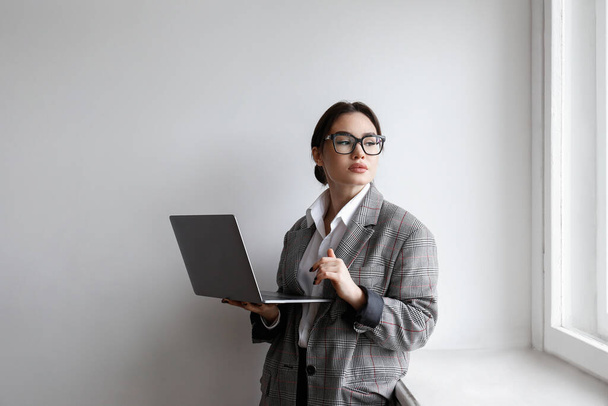 Portrait of a young businesswoman wearing smart casual attire standing with a laptop. Studio shot of a brunette female in eyeglasses posing near white wall. Close up, copy space, background. - Фото, изображение