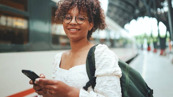 Close-up portrait of a young woman in glasses with a smartphone and backpack stands at the station and smiling. Positive woman using mobile phone outdoors in urban background. - Fotoğraf, Görsel