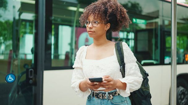 Young woman in glasses looking at a smartphone while standing at a bus stop. Positive woman using mobile phone outdoors in urban background. - Фото, изображение