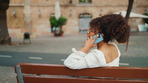 Young woman in glasses sits on a outdoor bench smiling and speaks on the smartphone. Positive woman using mobile phone outdoors in urban background. - 写真・画像
