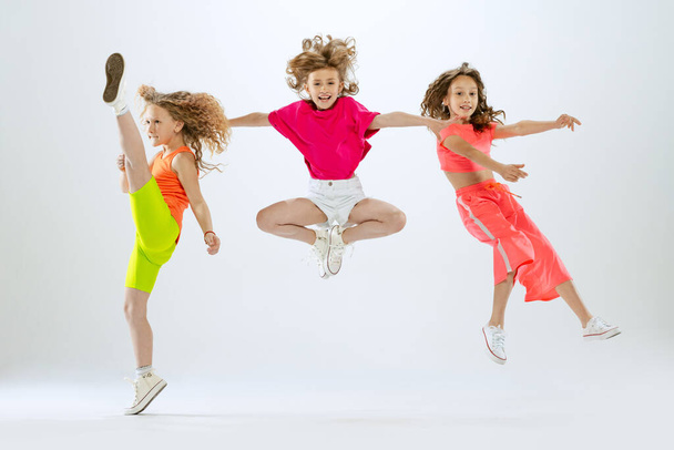 Dance group. Three little girls, kids in bright colorful clothes dancing, posing and jumping isolated on white studio background. Concept of music, fashion, art, childhood, hobby. Copy space for ad - Фото, изображение