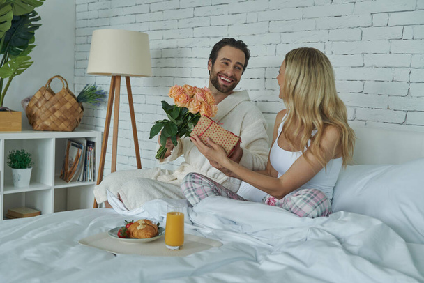 Cheerful man giving a gift box and flowers to his girlfriend while sitting in bed together - Foto, immagini