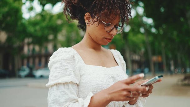 Young woman in glasses stand with a smartphone in her hands and looks around. Woman using mobile phone outdoors in urban background. - Fotoğraf, Görsel