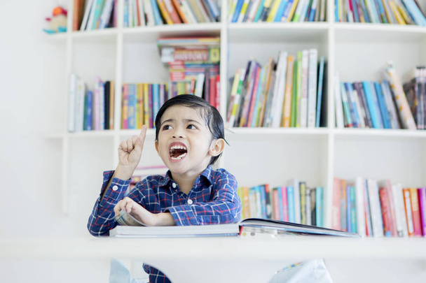 Cute little boy getting an idea while reading a book in the library with bookshelf background - Photo, Image