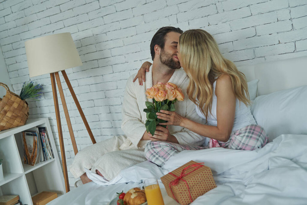 Young man kissing his girlfriend and giving a bunch of flowers while both sitting in bed - Foto, Bild