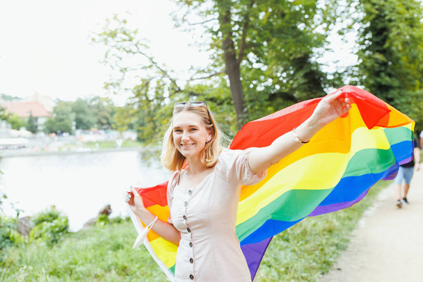 Happy young blonde hair woman covering with LGBTQI pride rainbow flag on the street in the park. LGBT Pride Month and Gay parade concept - Photo, Image