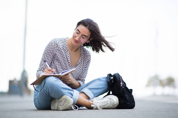 Portret van een jonge vrouw die buiten zit en notities in een boek schrijft - Foto, afbeelding