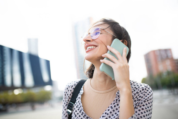 Close up portrait of beautiful young woman wearing eyeglasses looking away while talking on phone outside and smiling - Photo, Image