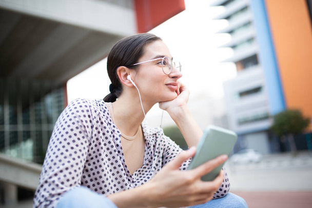 Retrato de cerca de una joven sentada afuera usando auriculares para escuchar música desde el teléfono móvil y mirando hacia otro lado con la mano en la barbilla - Foto, imagen