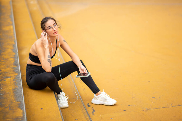 Portrait of fit young woman relaxing on steps outside with mobile phone and earphones listening to music after workout session - Foto, Imagem